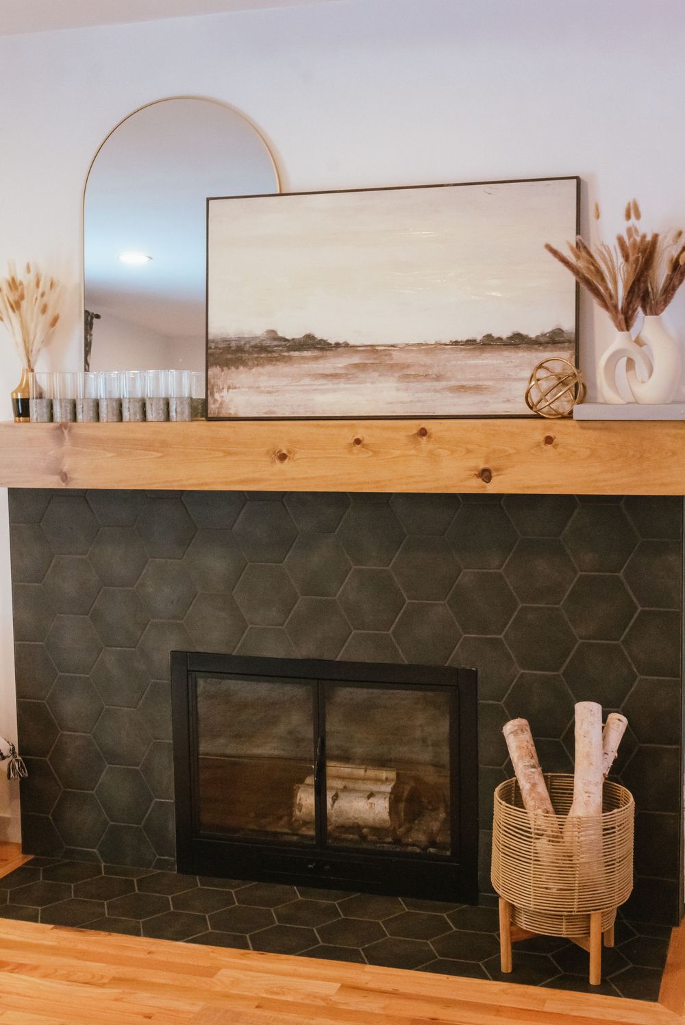 Modern farmhouse fireplace with black hexagon tile surround, arched mirror, and neutral decor on wooden mantel shelf.