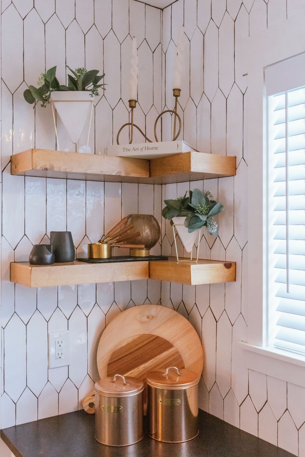 Modern kitchen corner with wooden floating shelves, geometric white tile wall, and copper canisters on dark countertop.