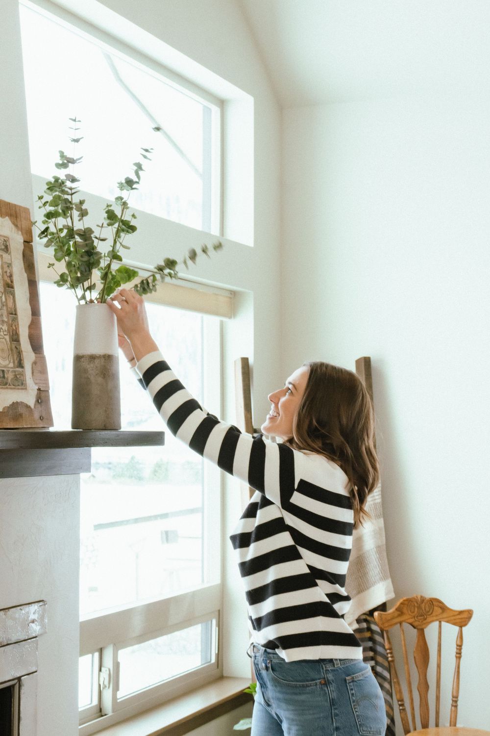 Person in striped sweater reaching up to adjust a plant on a shelf near a bright window.