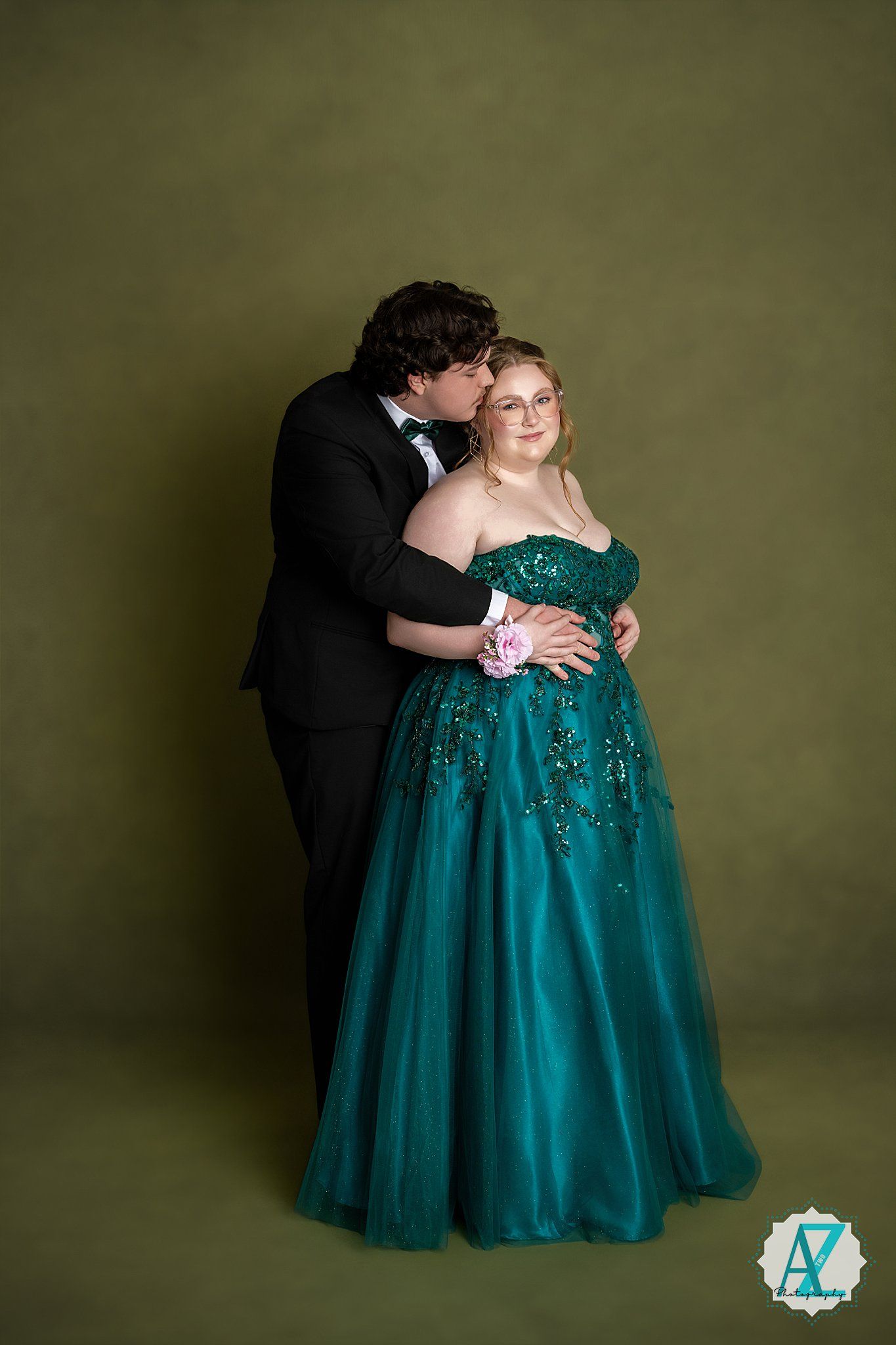 A couple embraces in formal wear with person in emerald sequined ball gown and pink corsage against olive backdrop.