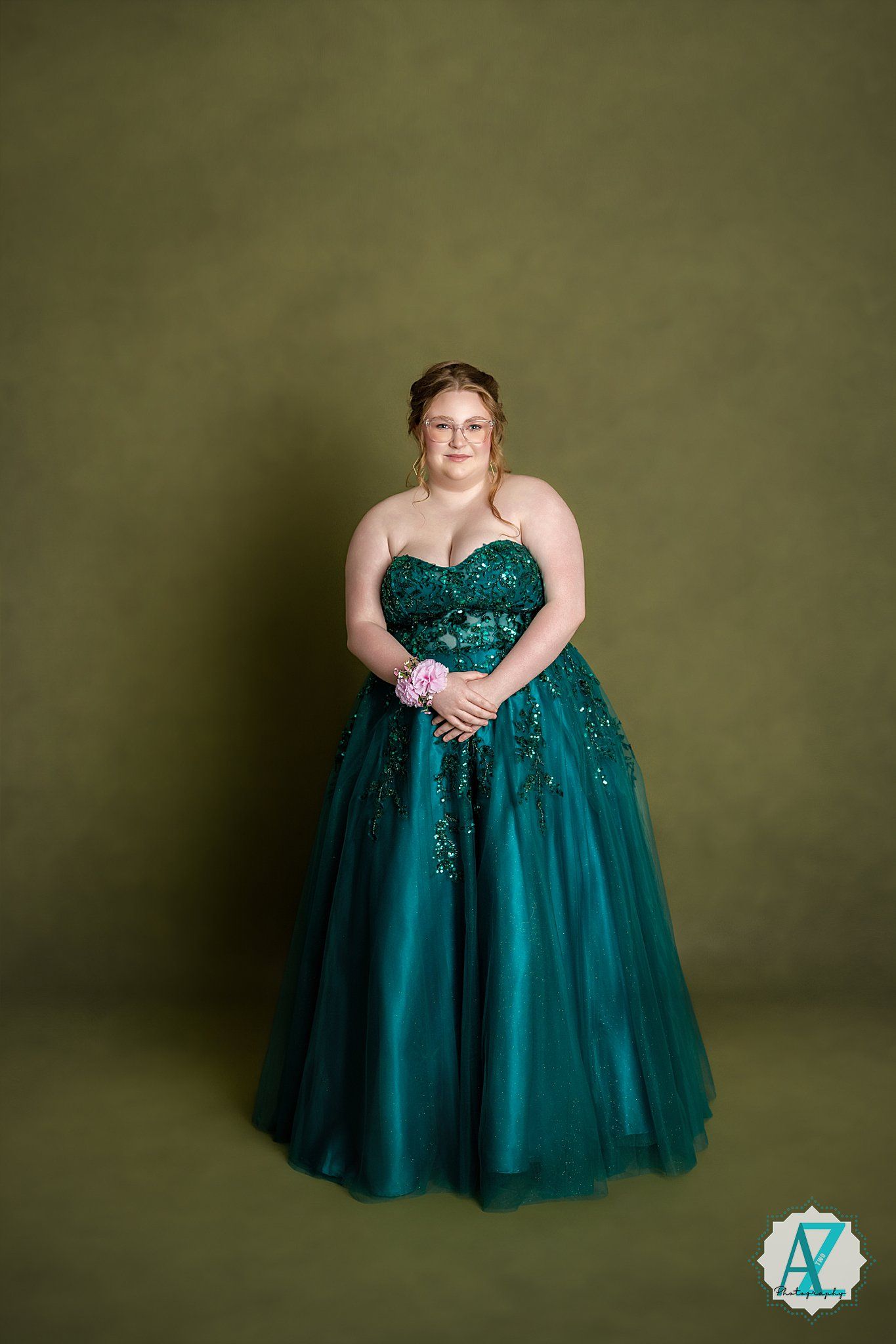 Someone wearing a stunning emerald green ball gown with sequins and tulle stands elegantly against an olive backdrop.