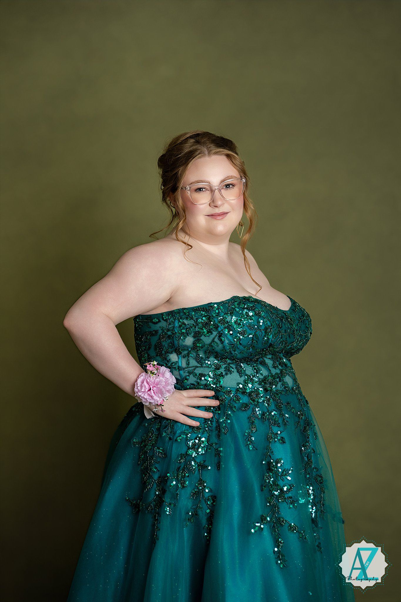 Person in emerald sequined ball gown with pink corsage poses against olive backdrop.