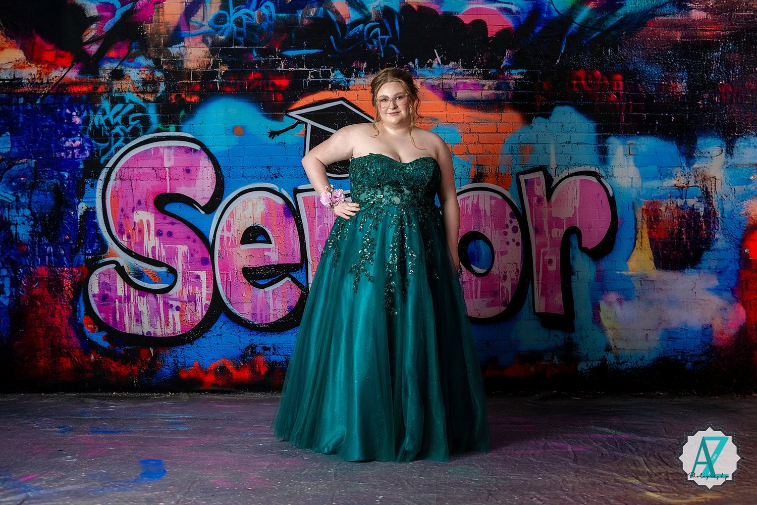 A person in a teal formal gown poses in front of colorful graffiti featuring the word 'Senior' during a photoshoot.