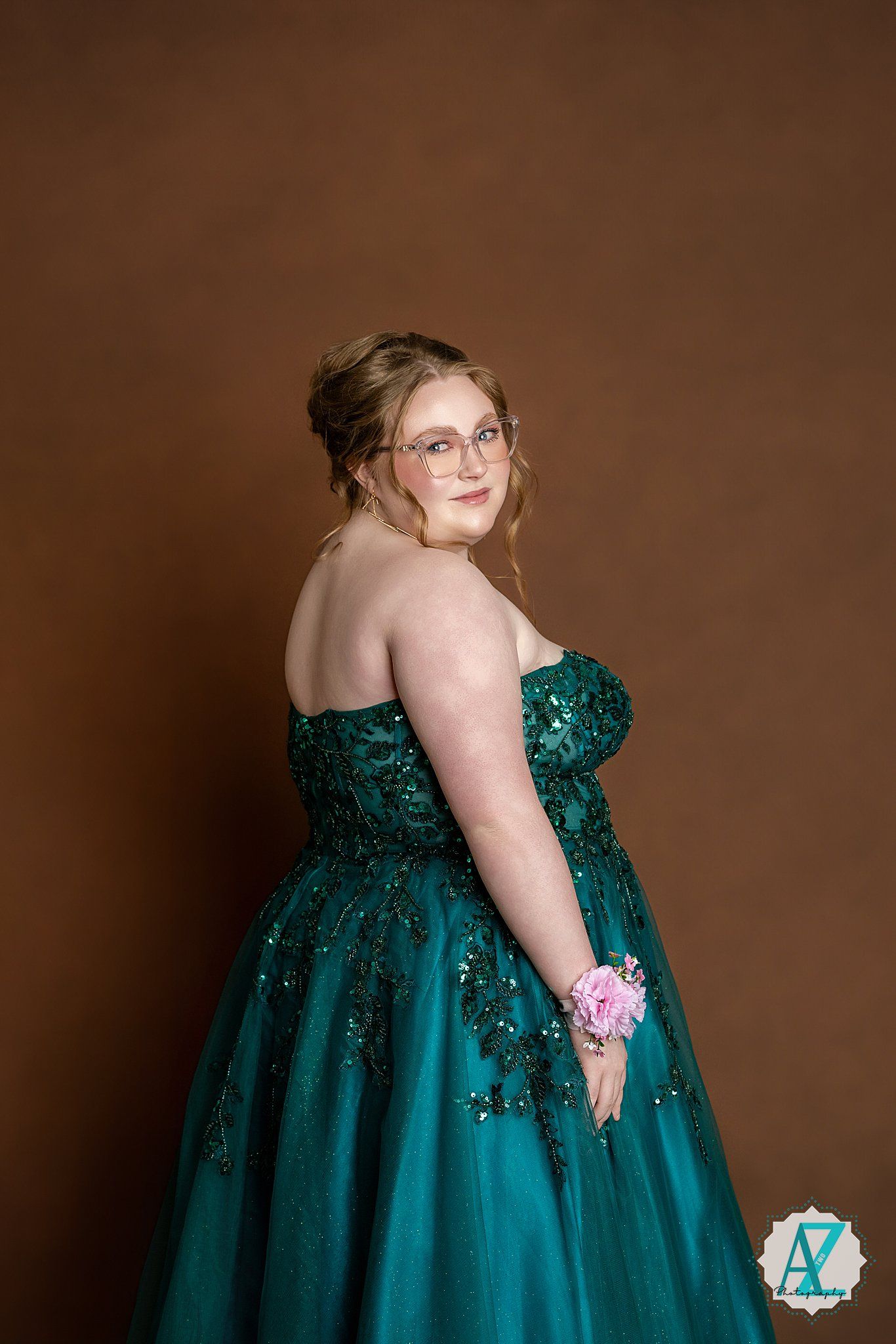 Person in teal sequined ball gown with pink corsage poses against brown backdrop.