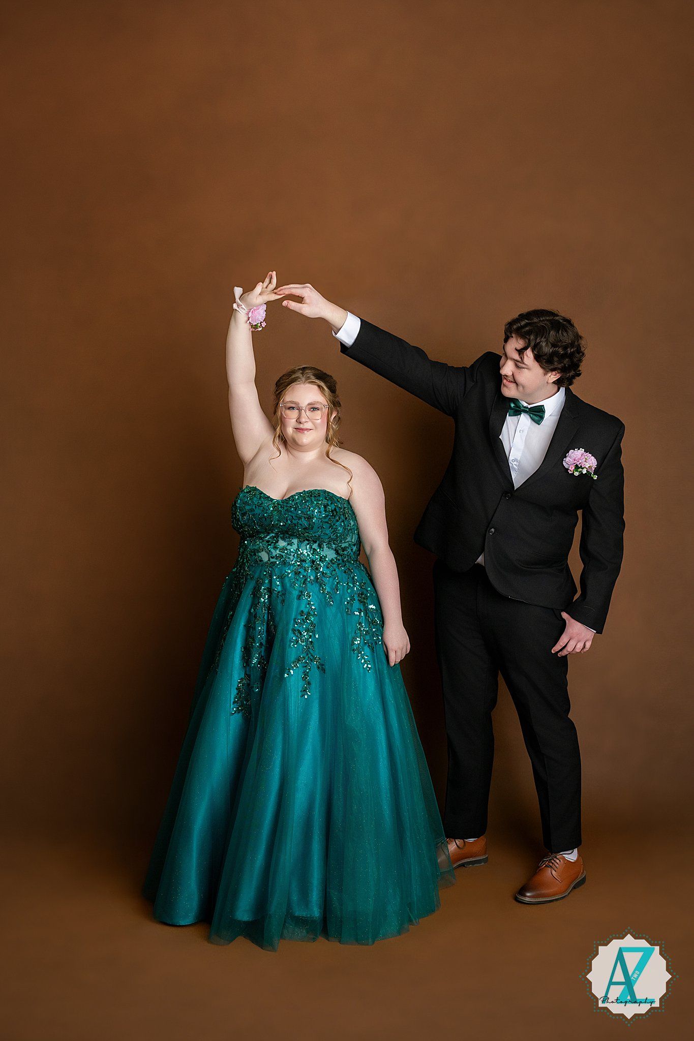 A couple in prom attire pose together, with a green sparkly ballgown and black tuxedo against a brown backdrop.