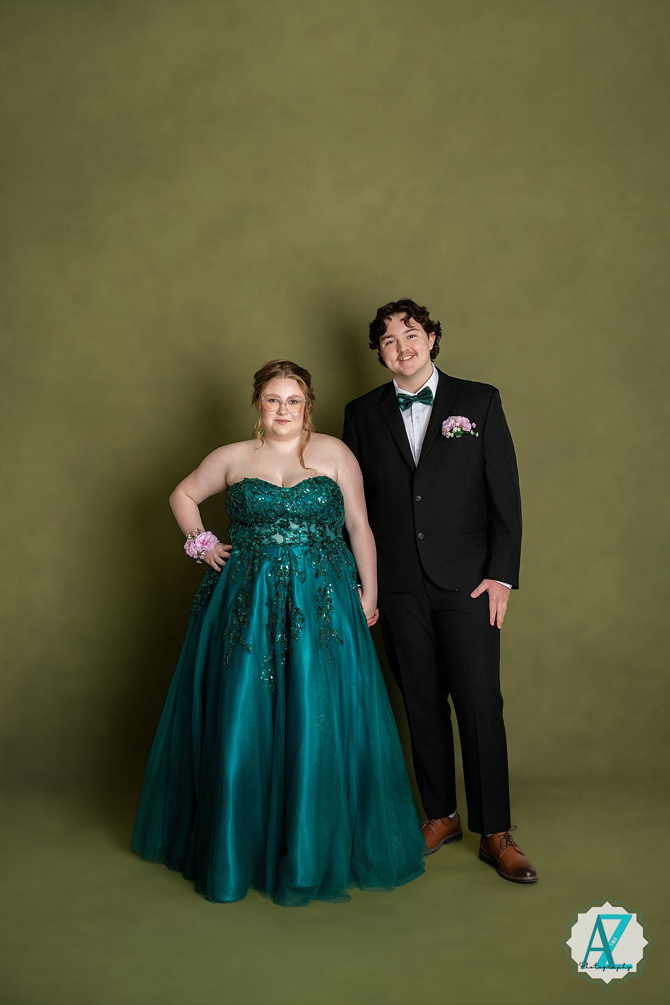 A formal couple poses against a green backdrop, with one in a stunning emerald sequined gown and the other in a black tuxedo.