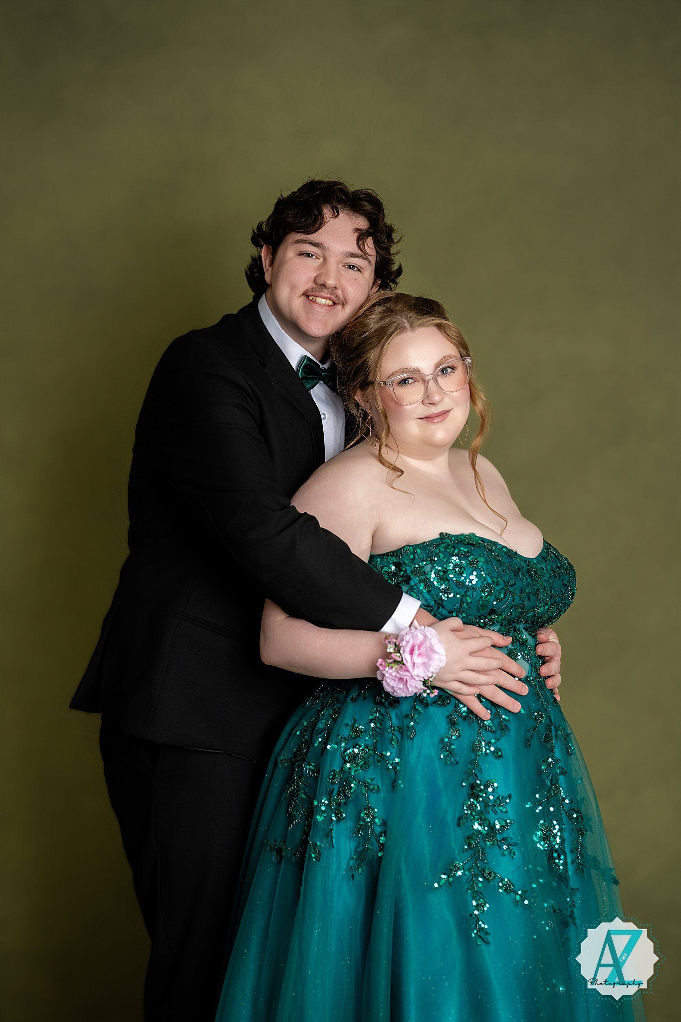 Couple in formal attire smiles together against olive backdrop wearing black tuxedo and emerald gown.