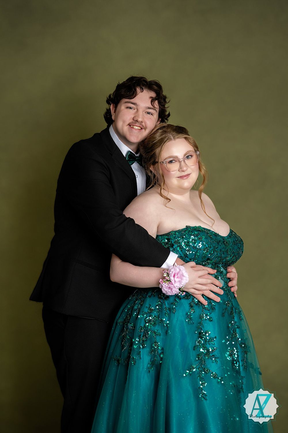 Couple in formal attire smiles together against olive backdrop wearing black tuxedo and emerald gown.
