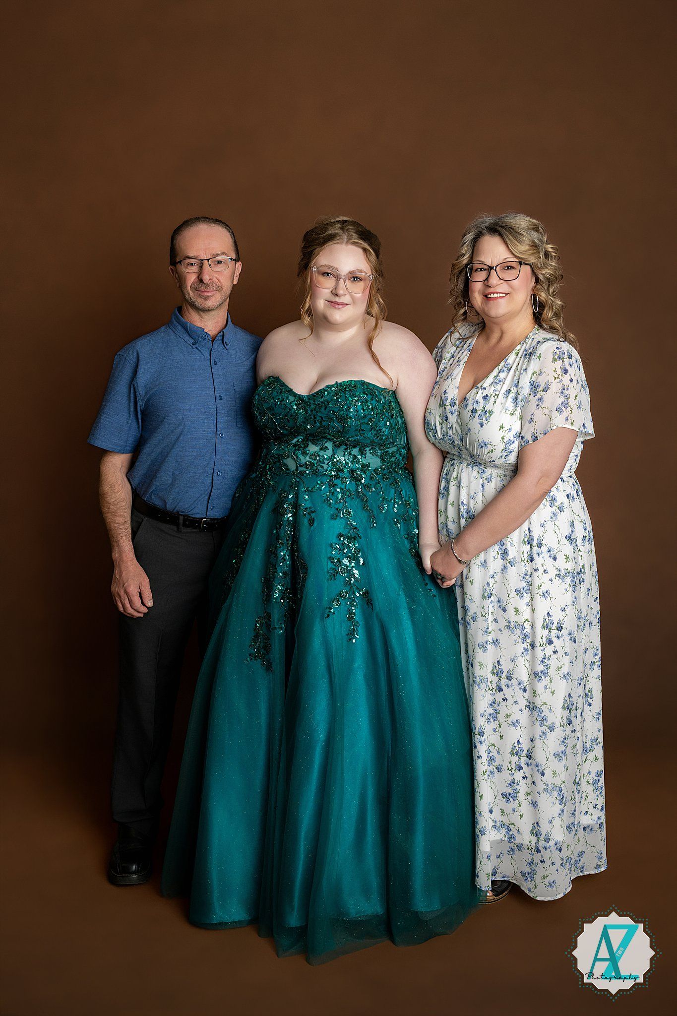 A formal portrait against a brown backdrop featuring three people in elegant attire including a stunning emerald green gown.