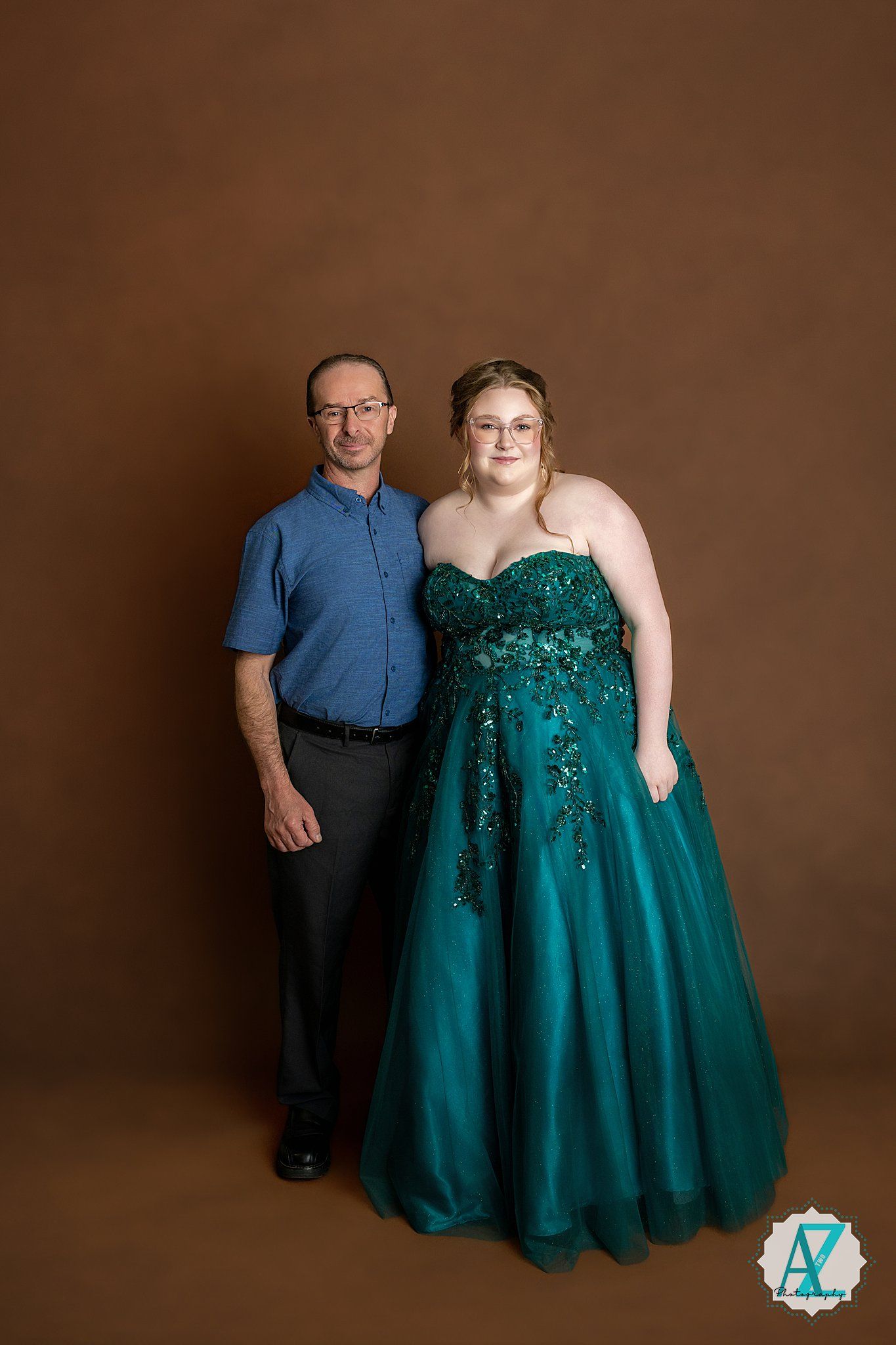 Two people pose in formal attire against a brown backdrop, with one wearing a sparkling emerald green ballgown.