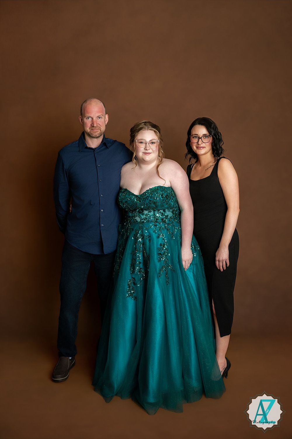 A formal portrait featuring three people against a brown backdrop with the center figure in a sparkly emerald gown.