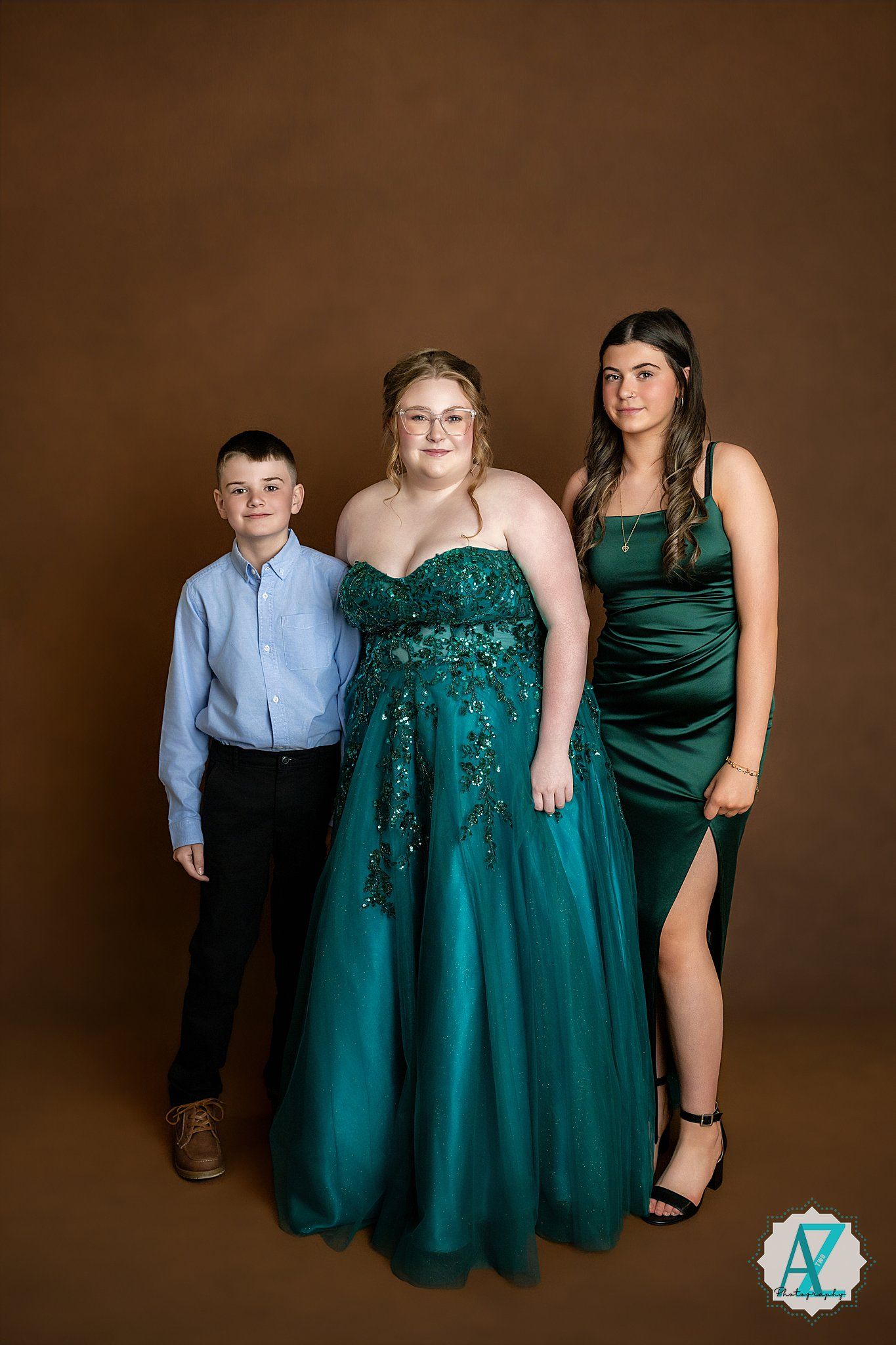 Studio portrait features three people in formal attire posing against a brown backdrop, wearing blue and emerald green outfits.