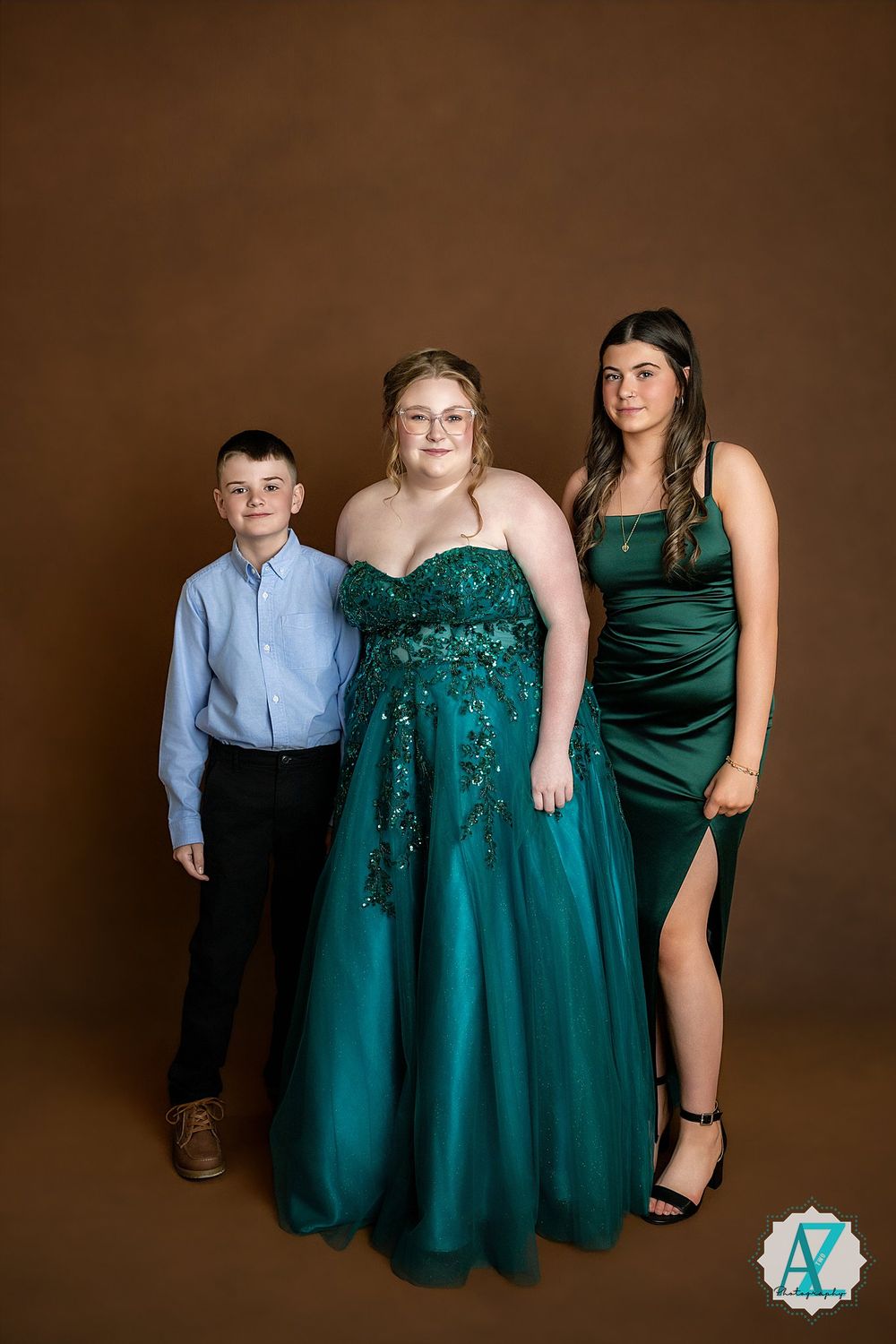 Studio portrait features three people in formal attire posing against a brown backdrop, wearing blue and emerald green outfits.