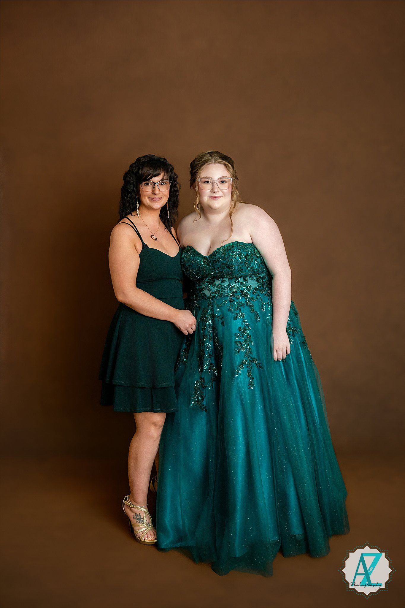 Two people in emerald green dresses pose for studio portrait against brown backdrop.