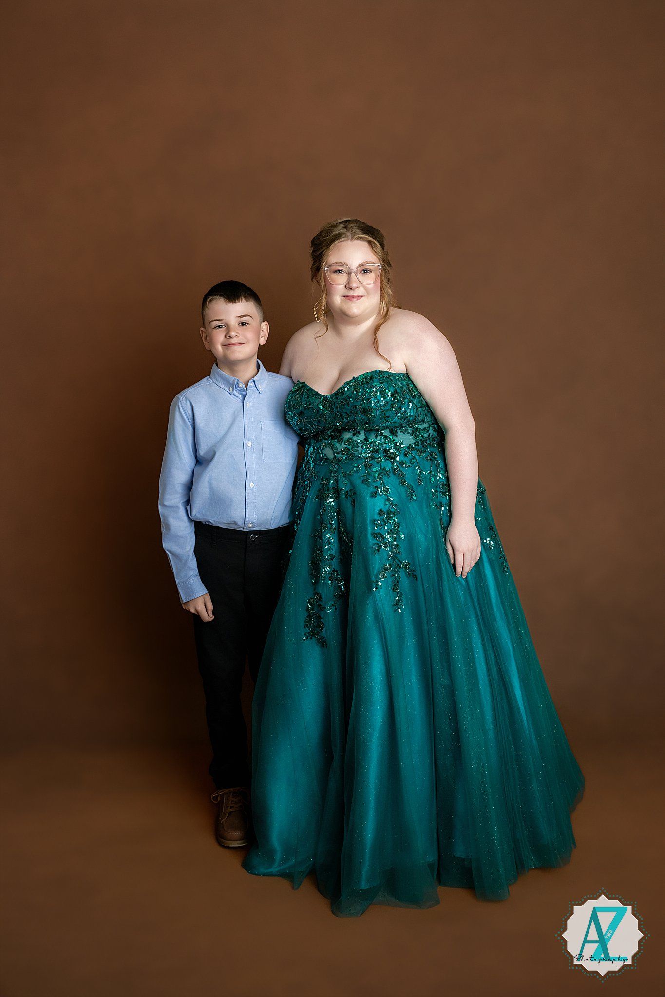 A formal portrait against brown backdrop shows two people posing together, with one wearing a sparkly emerald green ballgown.