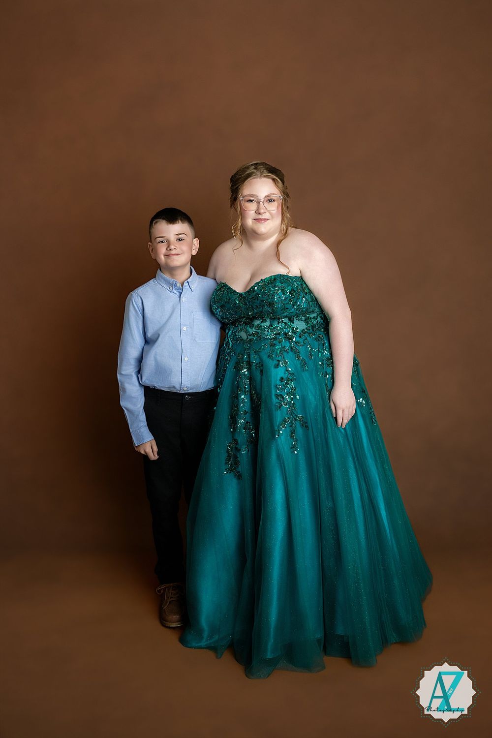A formal portrait against brown backdrop shows two people posing together, with one wearing a sparkly emerald green ballgown.