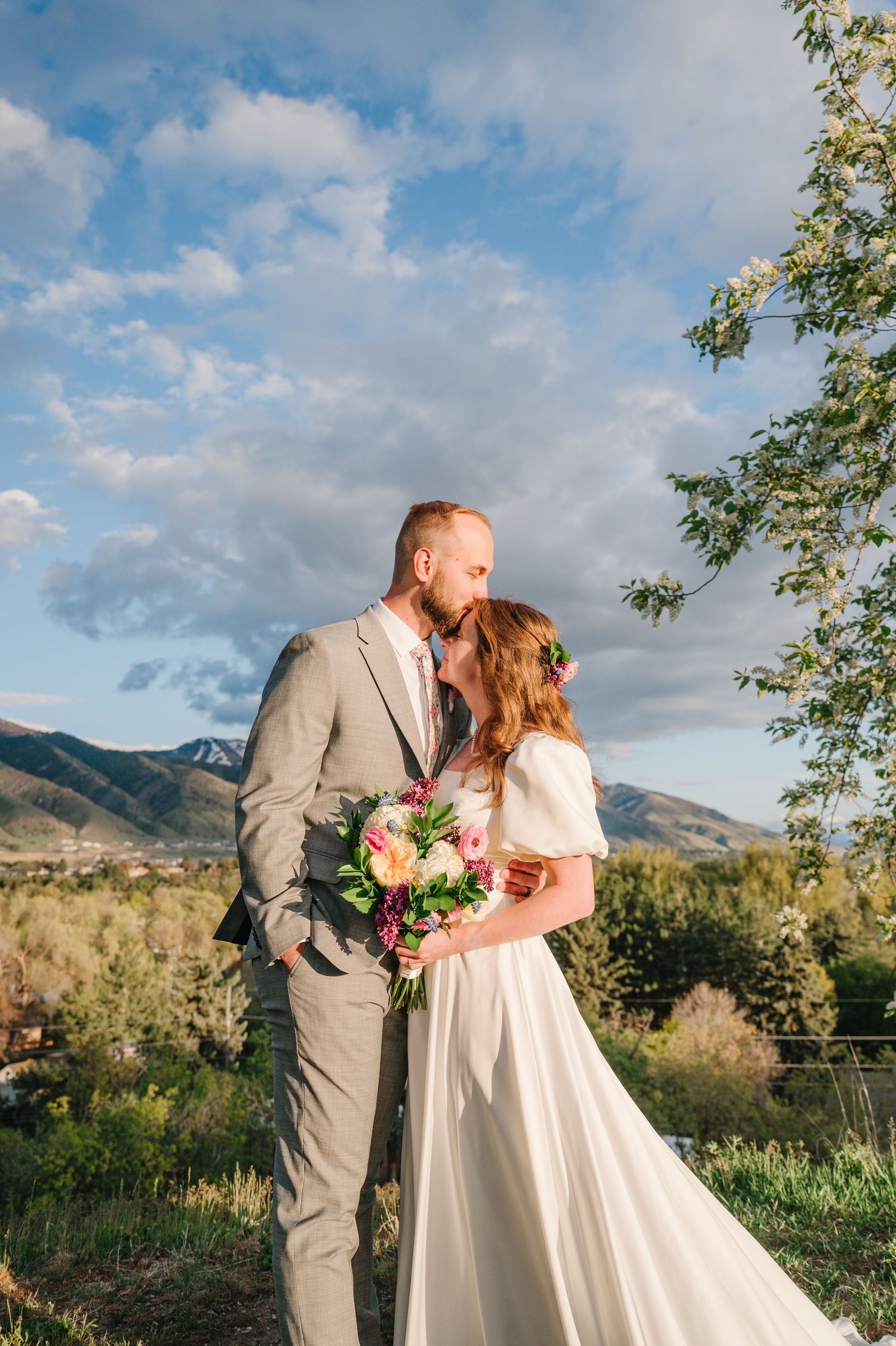 A couple in wedding attire embrace against a scenic mountain landscape with a bouquet of pink flowers at sunset.