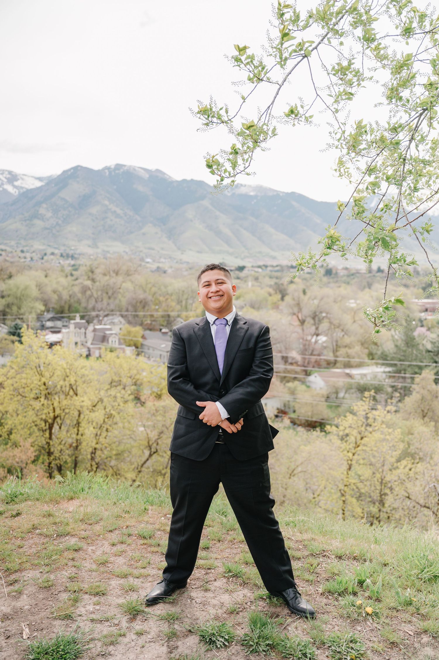 A person in a dark business suit smiles while standing on a hillside with mountains and spring foliage in the background.