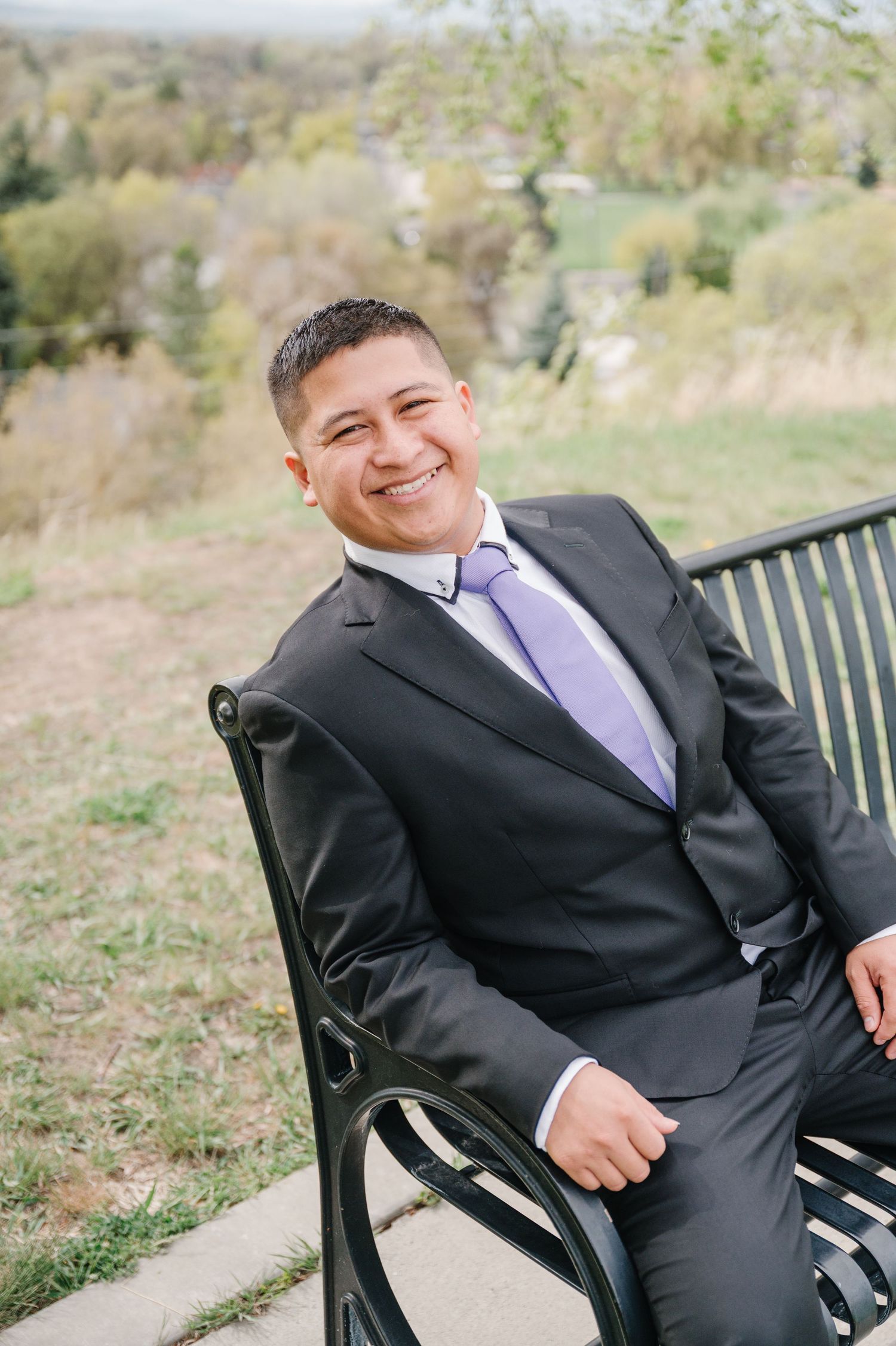 Professional portrait of someone in a dark suit and purple tie sitting casually on a park bench outdoors.