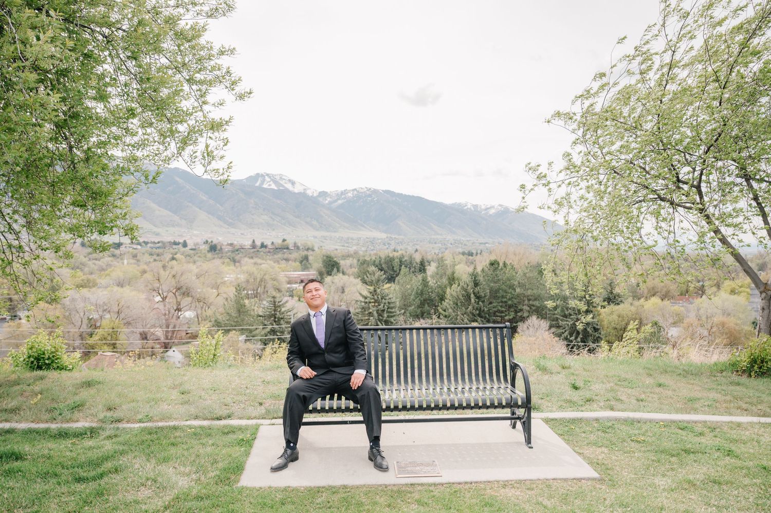 A person sits on a metal bench overlooking scenic mountain views with lush greenery and trees in the background.