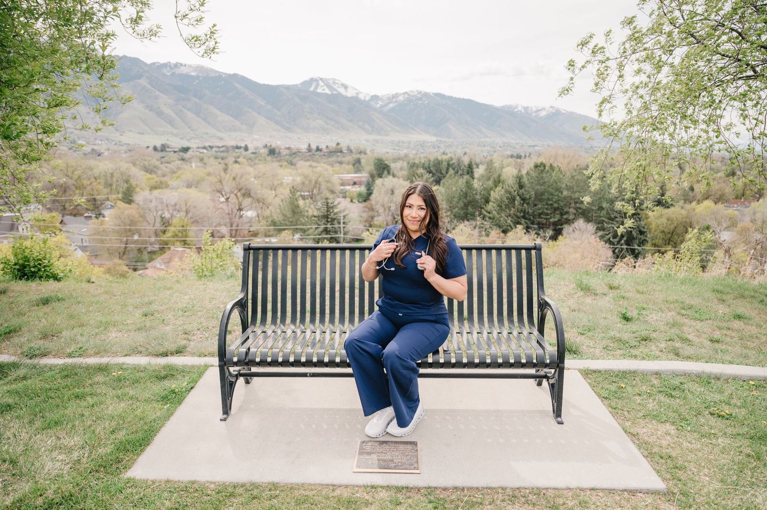 Someone sits relaxed on a metal bench with mountains and trees in the scenic background on a spring day.