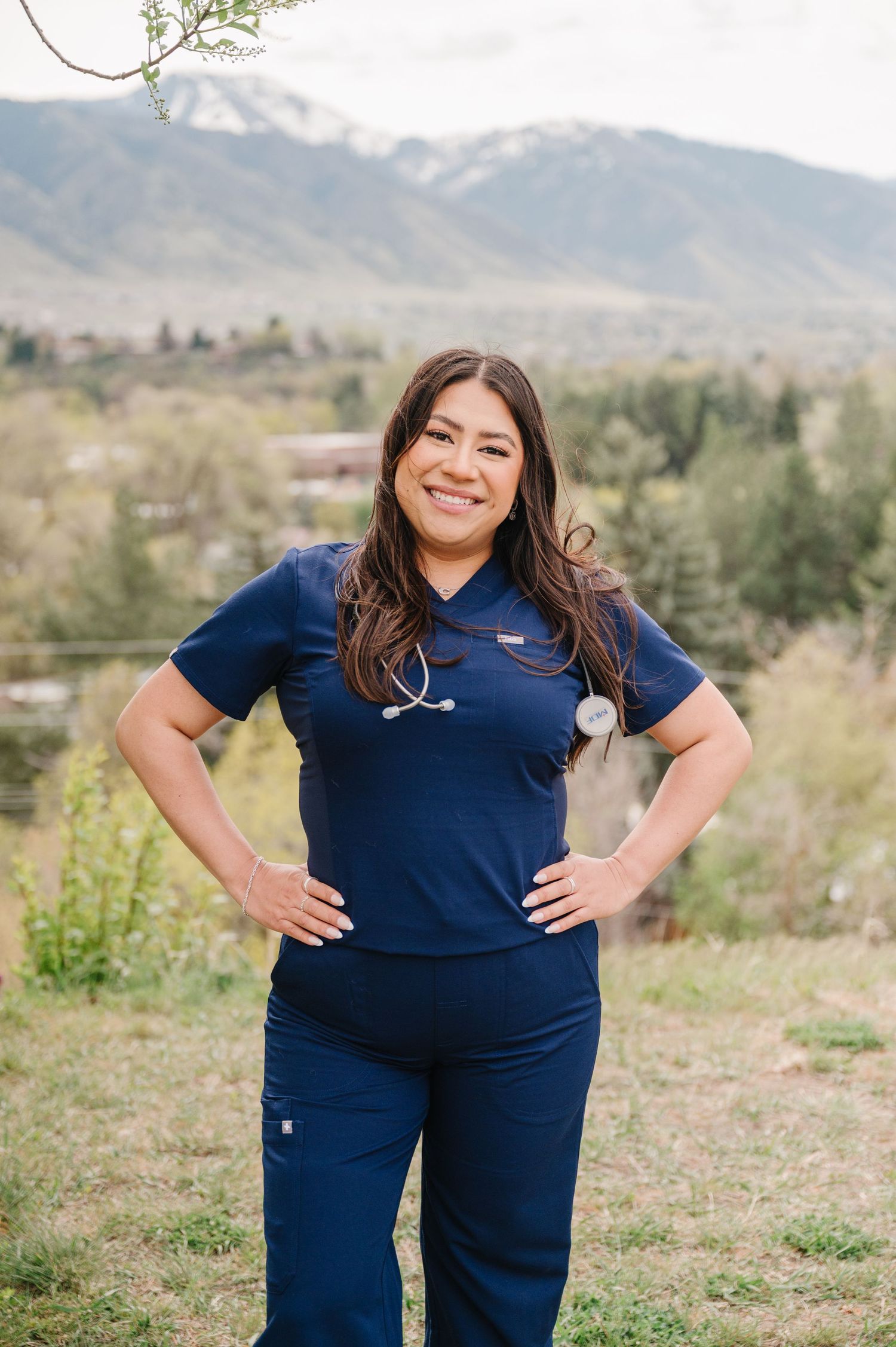 A medical professional in navy blue scrubs and stethoscope poses outdoors against a mountain backdrop.