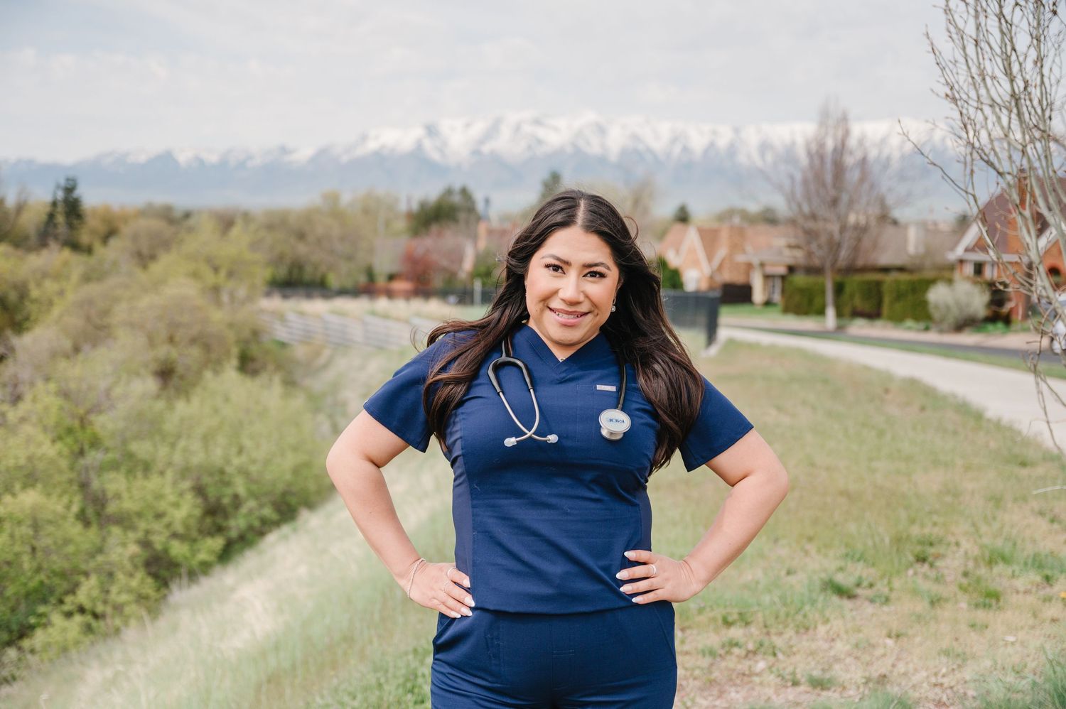 Healthcare professional in navy blue scrubs and stethoscope stands outside with snow-capped mountains in background.