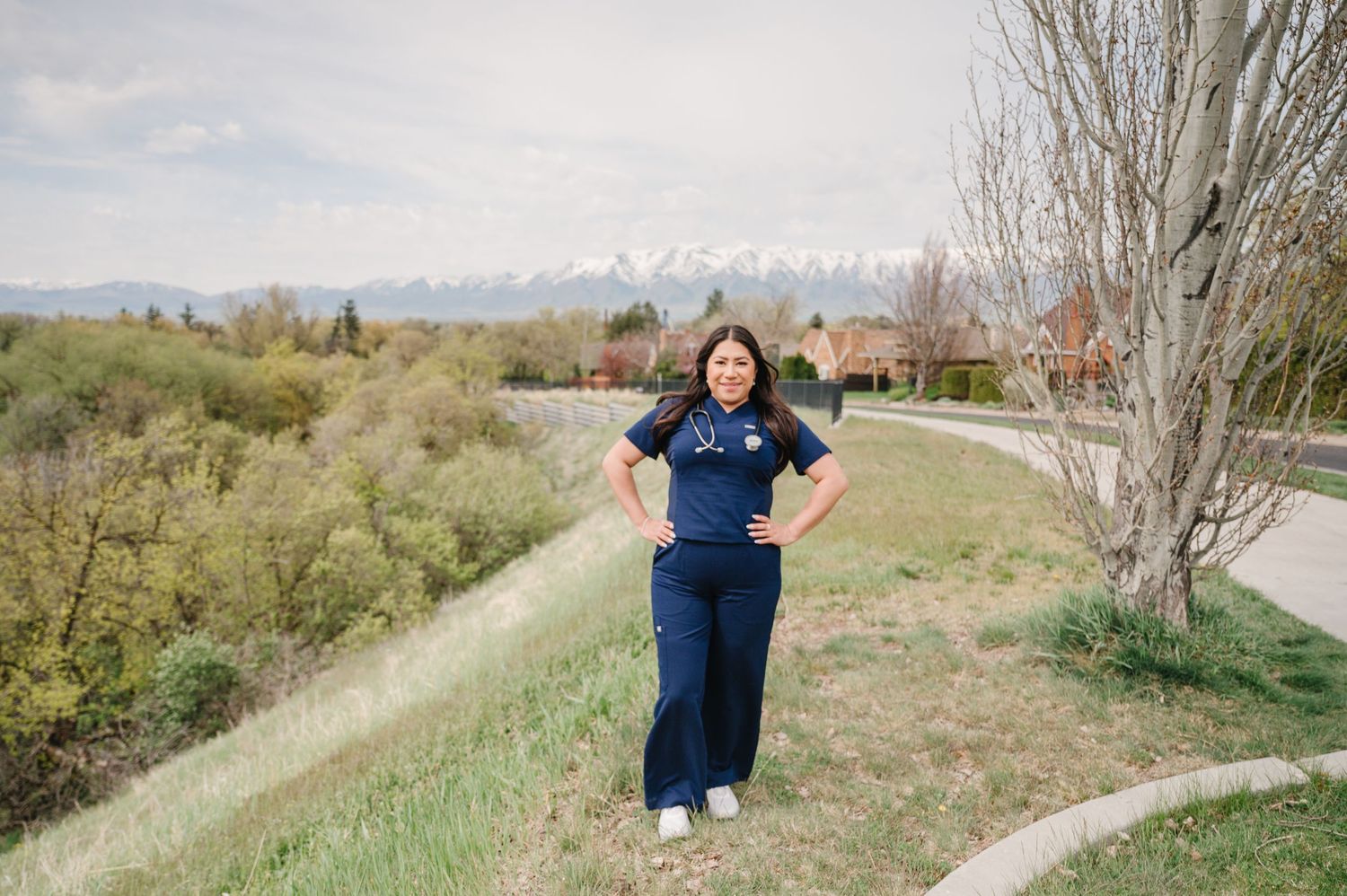 Someone posing on a grassy hillside path with mountains and bare trees visible in the background.