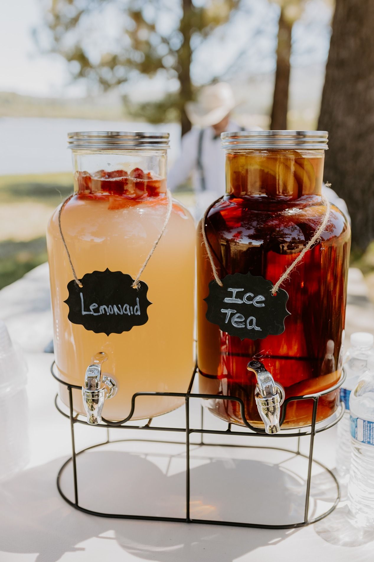 Two glass drink dispensers with lemonade and iced tea on a metal stand at an outdoor event.