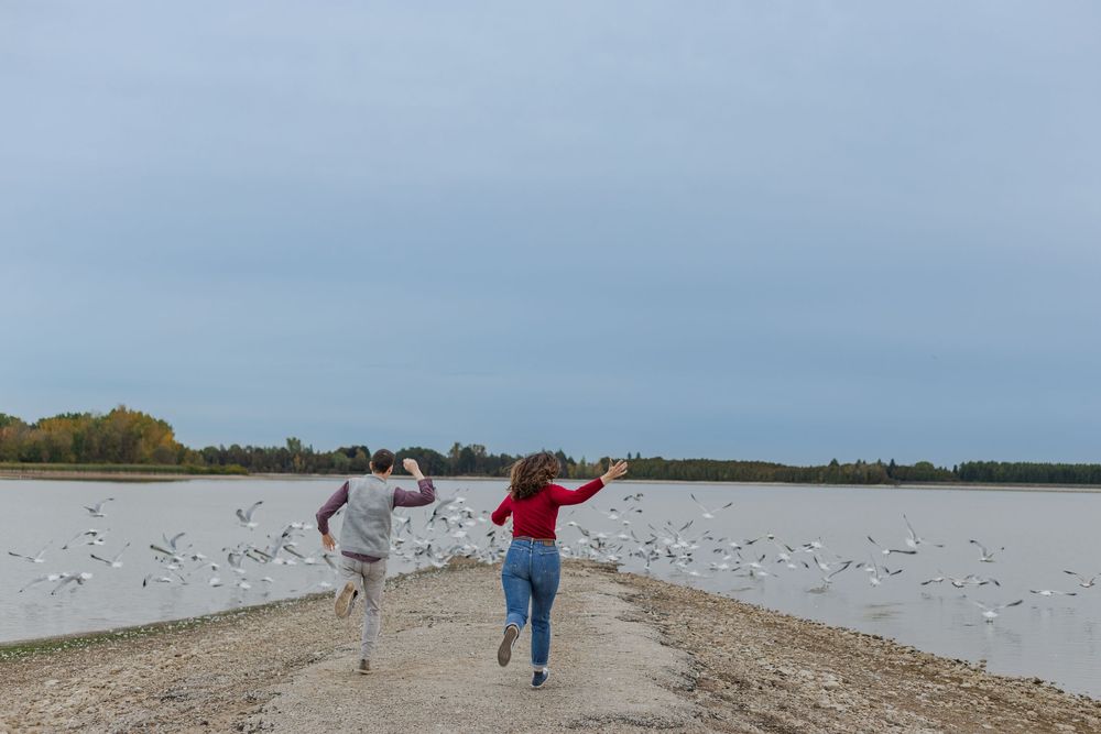 Playful engagement photo of couple chasing after birds by the water at the guelph lake conservation area in guelph, ontario.