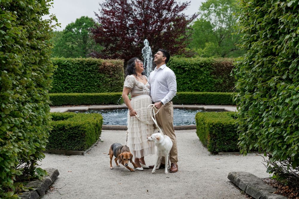 Natural engagement photo of couple standing with their dogs in front of the fountain at the guelph arboretum in guelph, ontario.