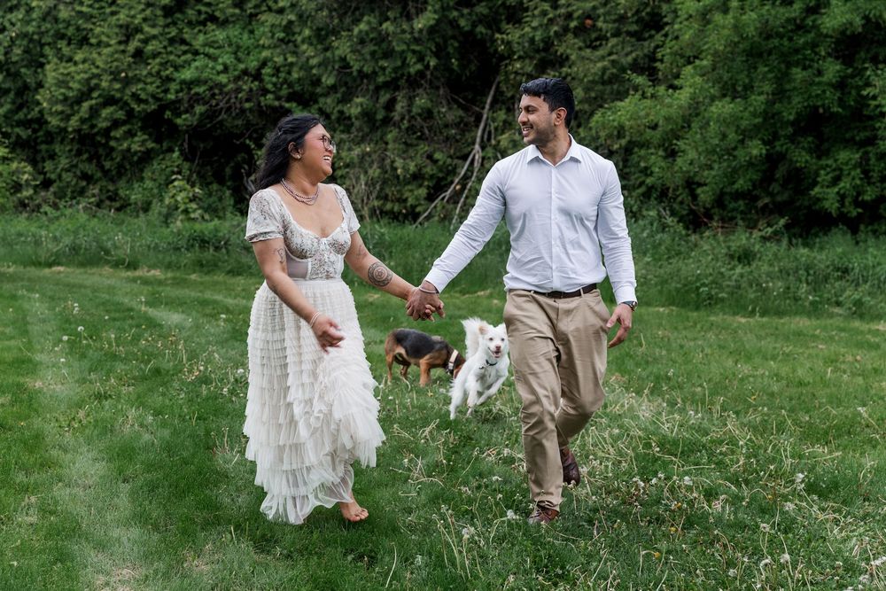 Candid engagement photo of couple running through a field with their dogs at the guelph arboretum in guelph, ontario.