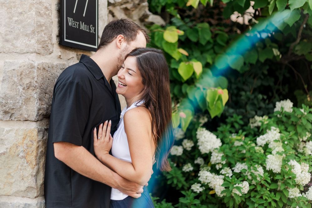 Candid engagement photo of couple leaning on stone wall at elora mill while laughing in downtown Elora, Ontario.