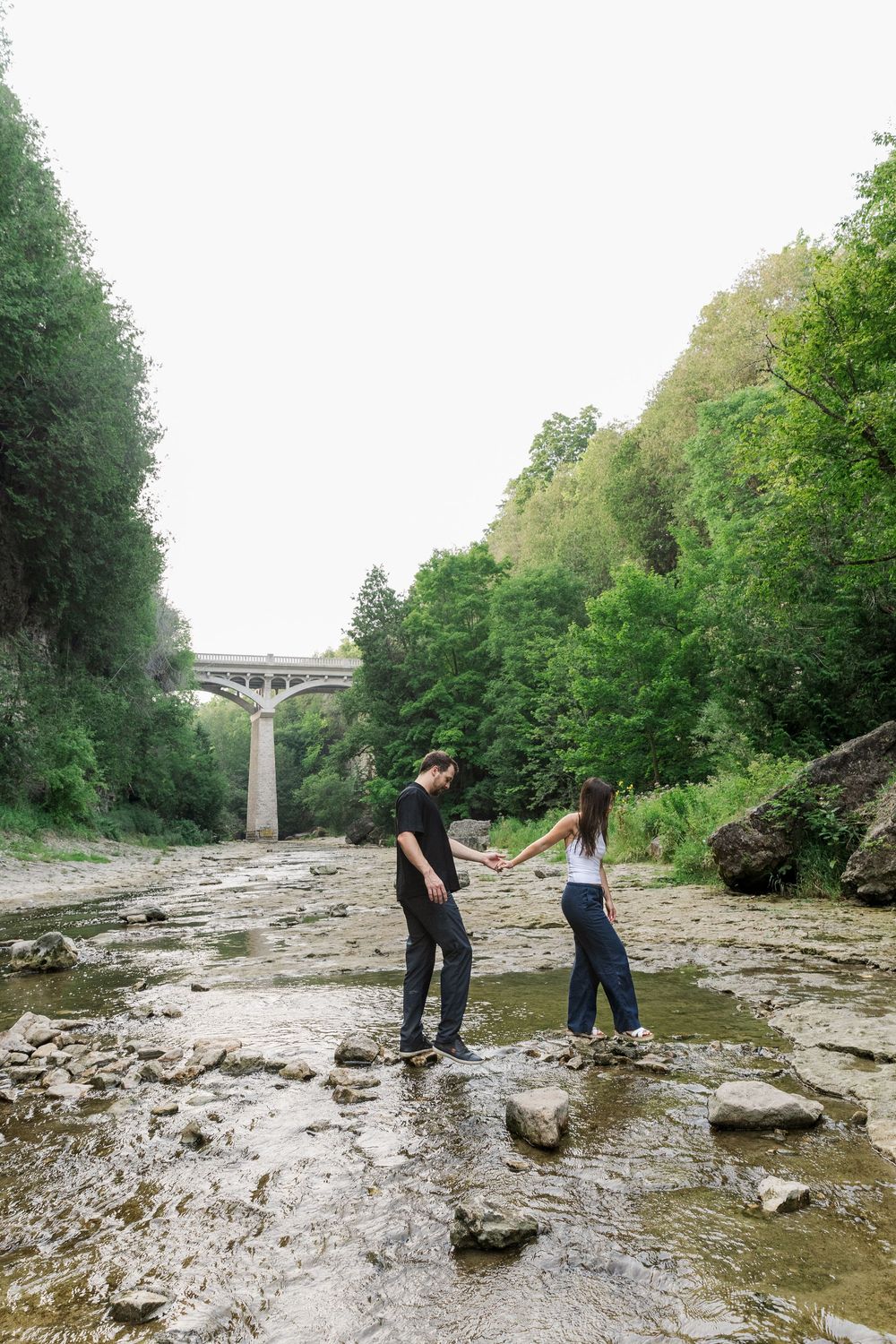 Couple walking together across the rocks on the river bed of the Elora gorge during their engagement session in Elora, Ontario.
