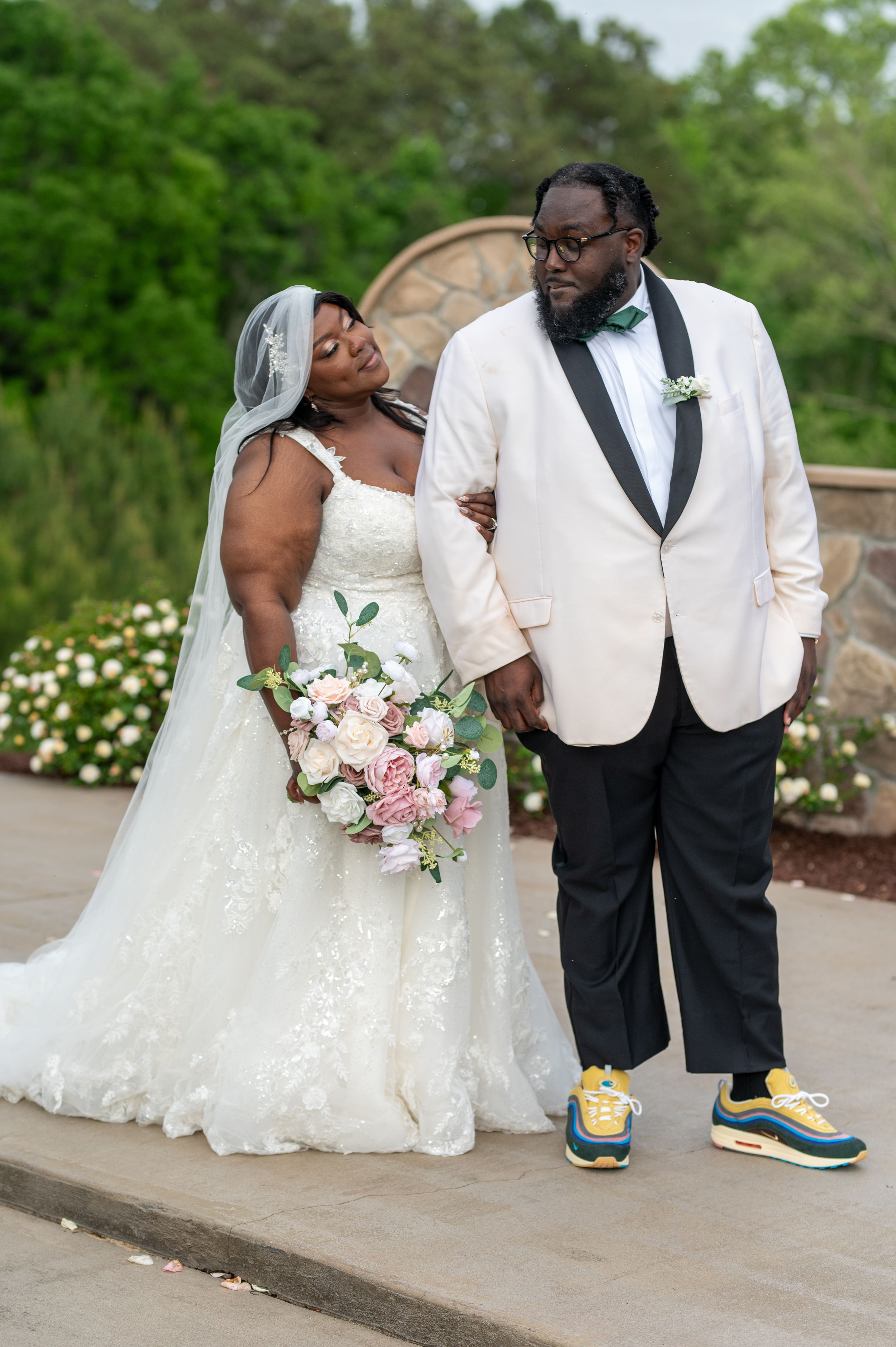 Charlotte photographer. Bride in white lace gown holds pink bouquet beside groom in ivory tuxedo and colorful sneakers at outdoor wedding.