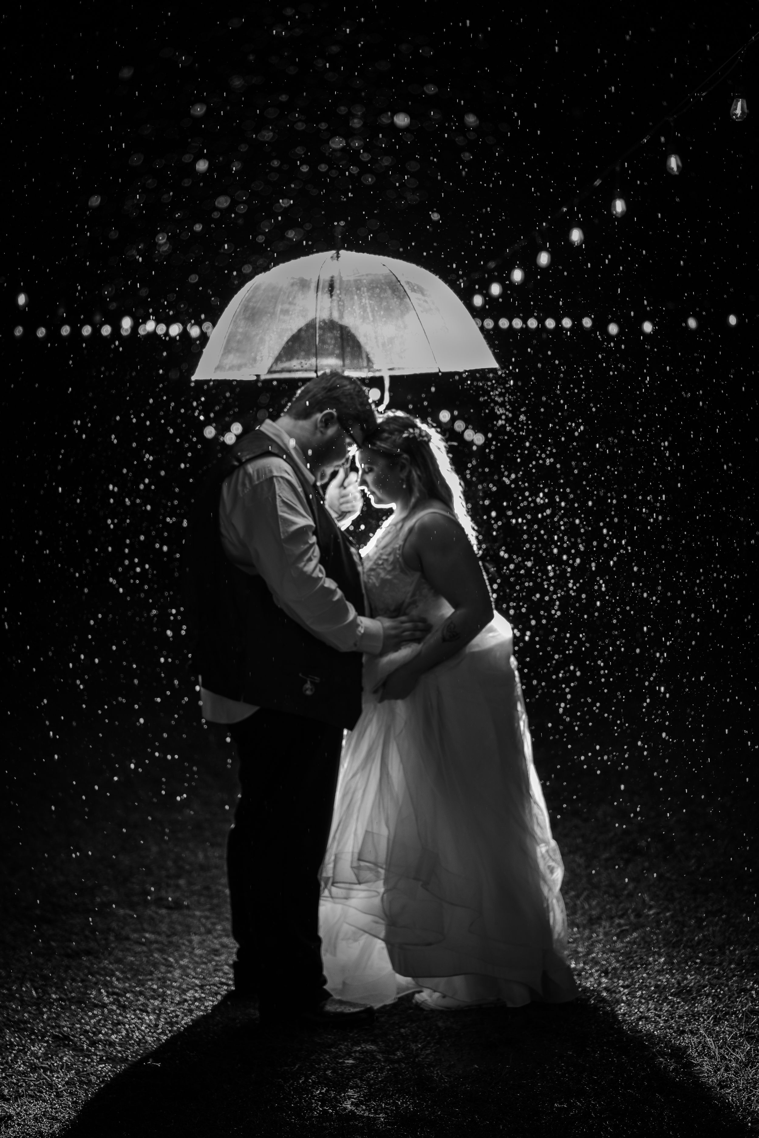 Black and white photo of bride and groom embracing under a clear umbrella in the rain at night. Charlotte wedding photographer.