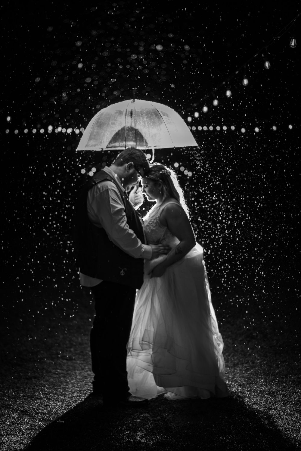 Black and white photo of bride and groom embracing under a clear umbrella in the rain at night. Charlotte wedding photographer.