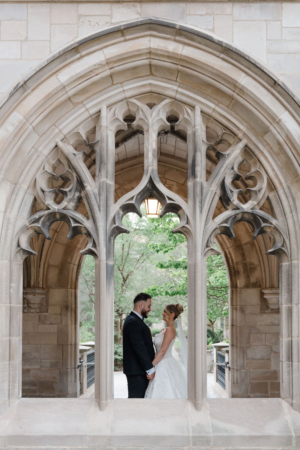 Bride and groom share a romantic moment framed by an ornate Gothic stone archway with lush greenery backdrop.