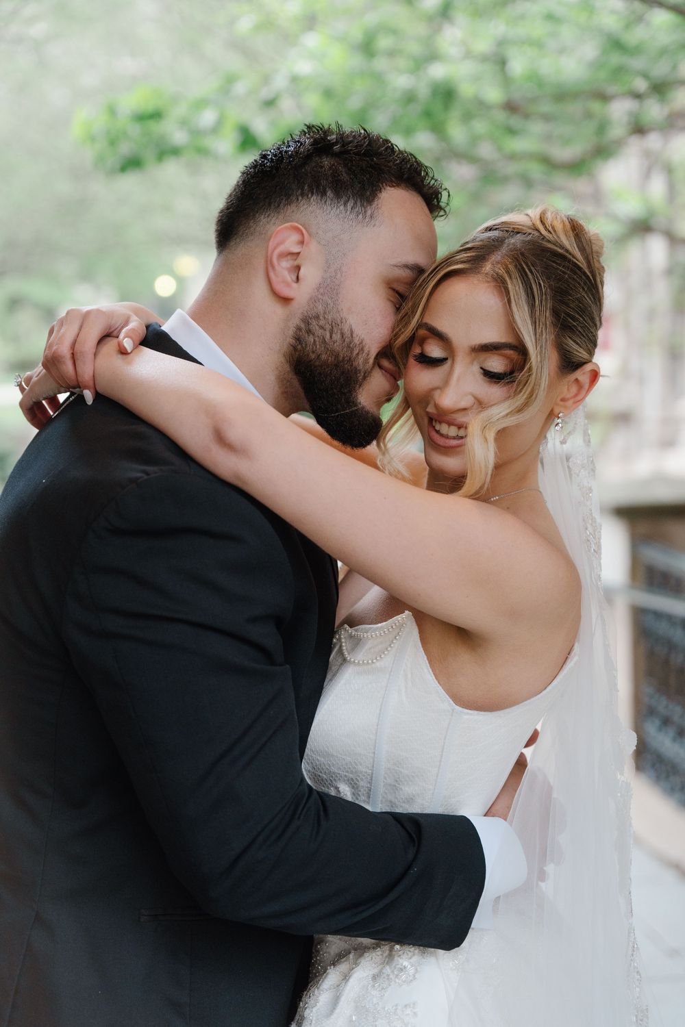 Bride and groom share a tender embrace outdoors, smiling joyfully in wedding attire among lush green trees.