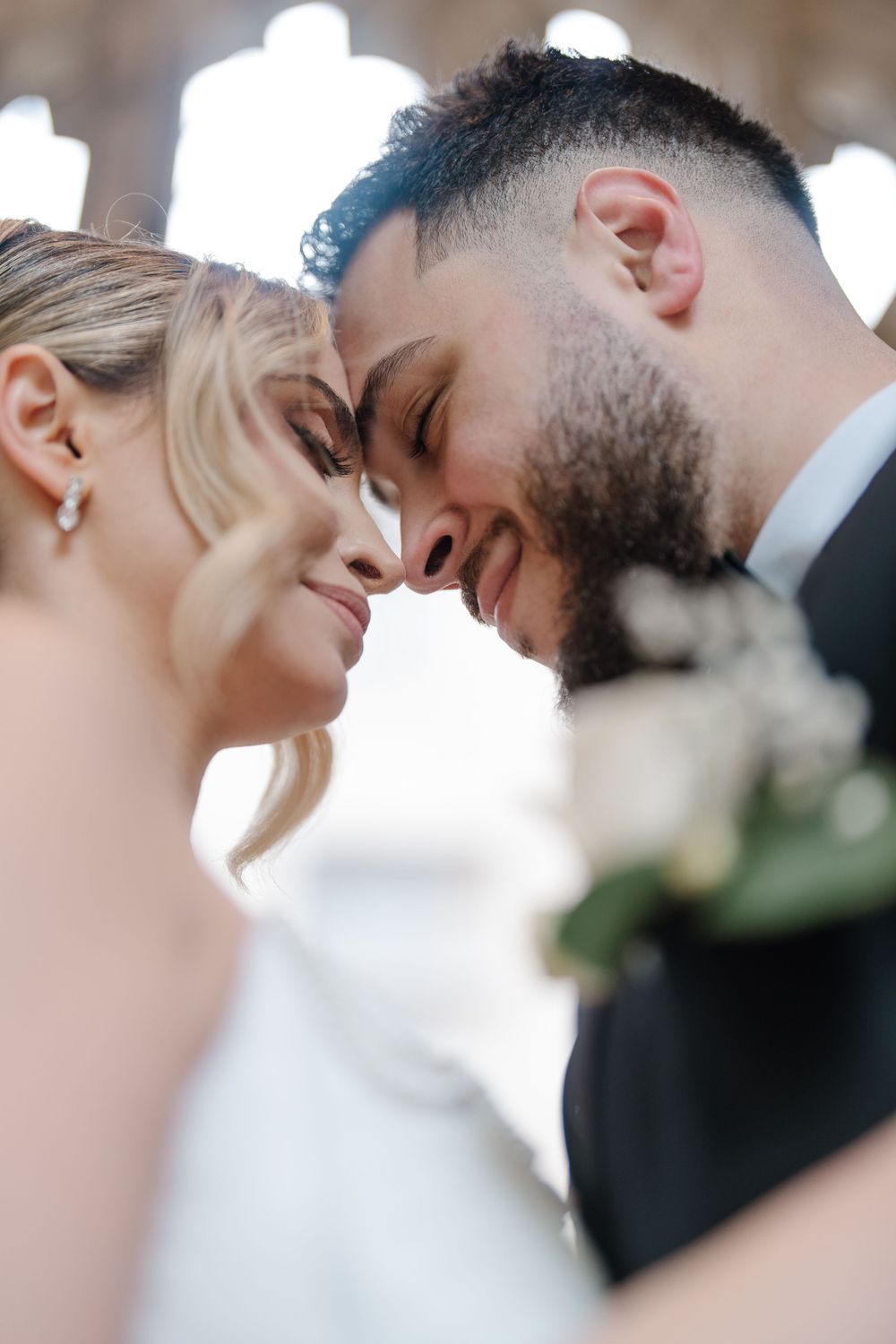 Smiling bride and groom touch foreheads intimately, bride holding white roses bouquet in soft light.