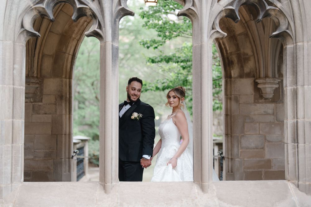 Bride and groom hold hands and smile while posing beneath elegant Gothic stone arches outdoors.