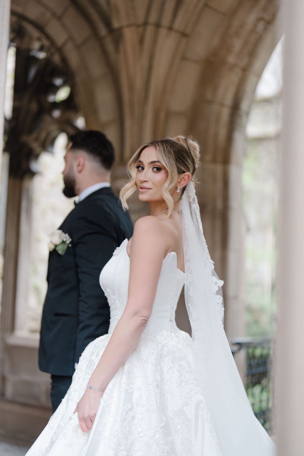 Bride in white lace ballgown with veil smiles over shoulder beside groom in dark suit under stone arches.