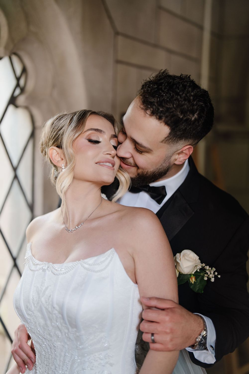 Groom in black tuxedo tenderly kisses bride in strapless lace gown near ornate stone architecture at wedding.