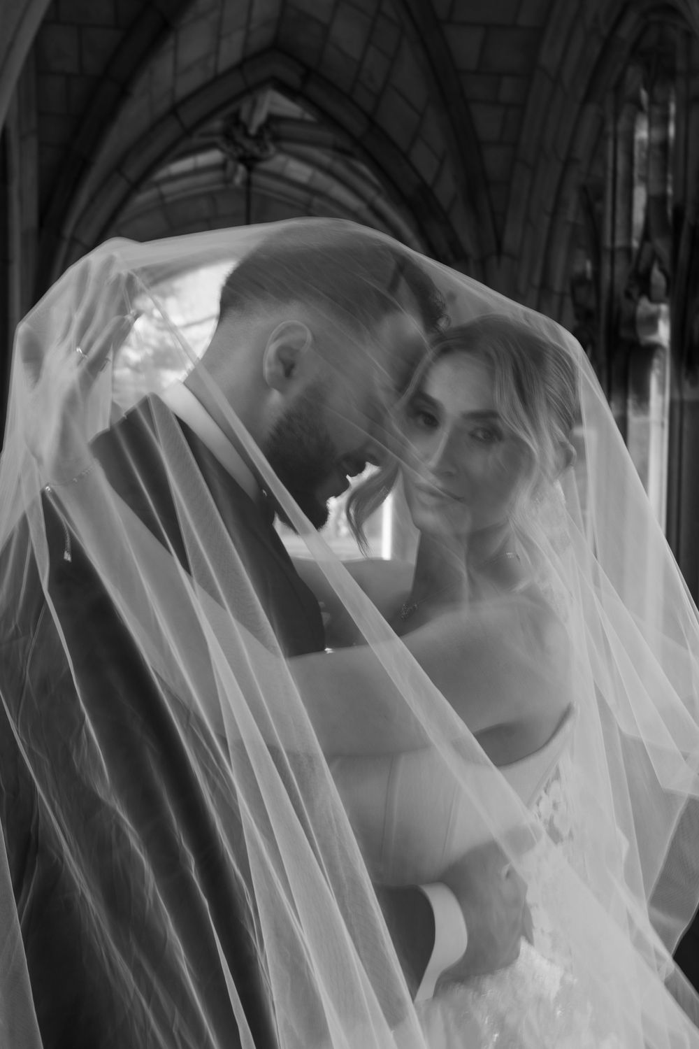 Black and white wedding photo of bride and groom sharing intimate moment under veil in gothic church.