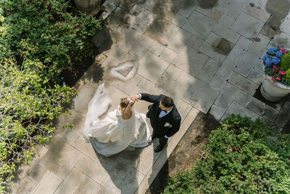 Aerial view of bride in white gown and groom dancing on stone courtyard surrounded by greenery.