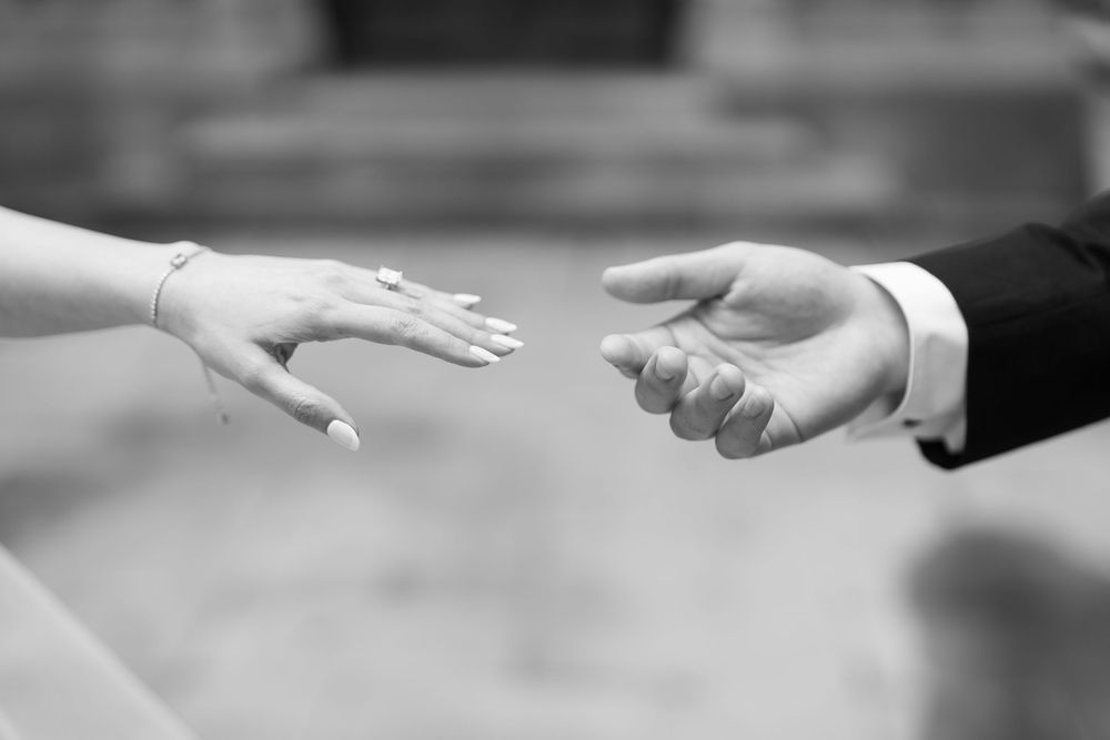 Black and white close-up of bride and groom reaching toward each other with outstretched hands.