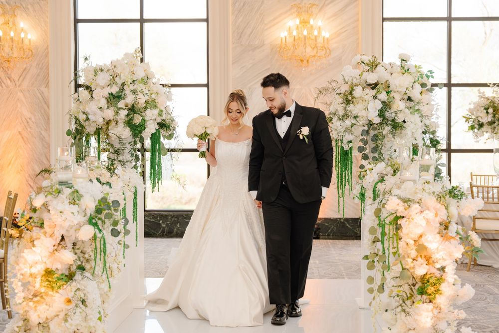 Bride and groom walk together under lush white floral arch with greenery at elegant wedding ceremony.