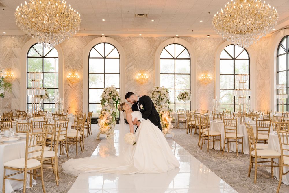 Bride and groom sharing a kiss on a white dance floor in a luxurious ballroom with crystal chandeliers.