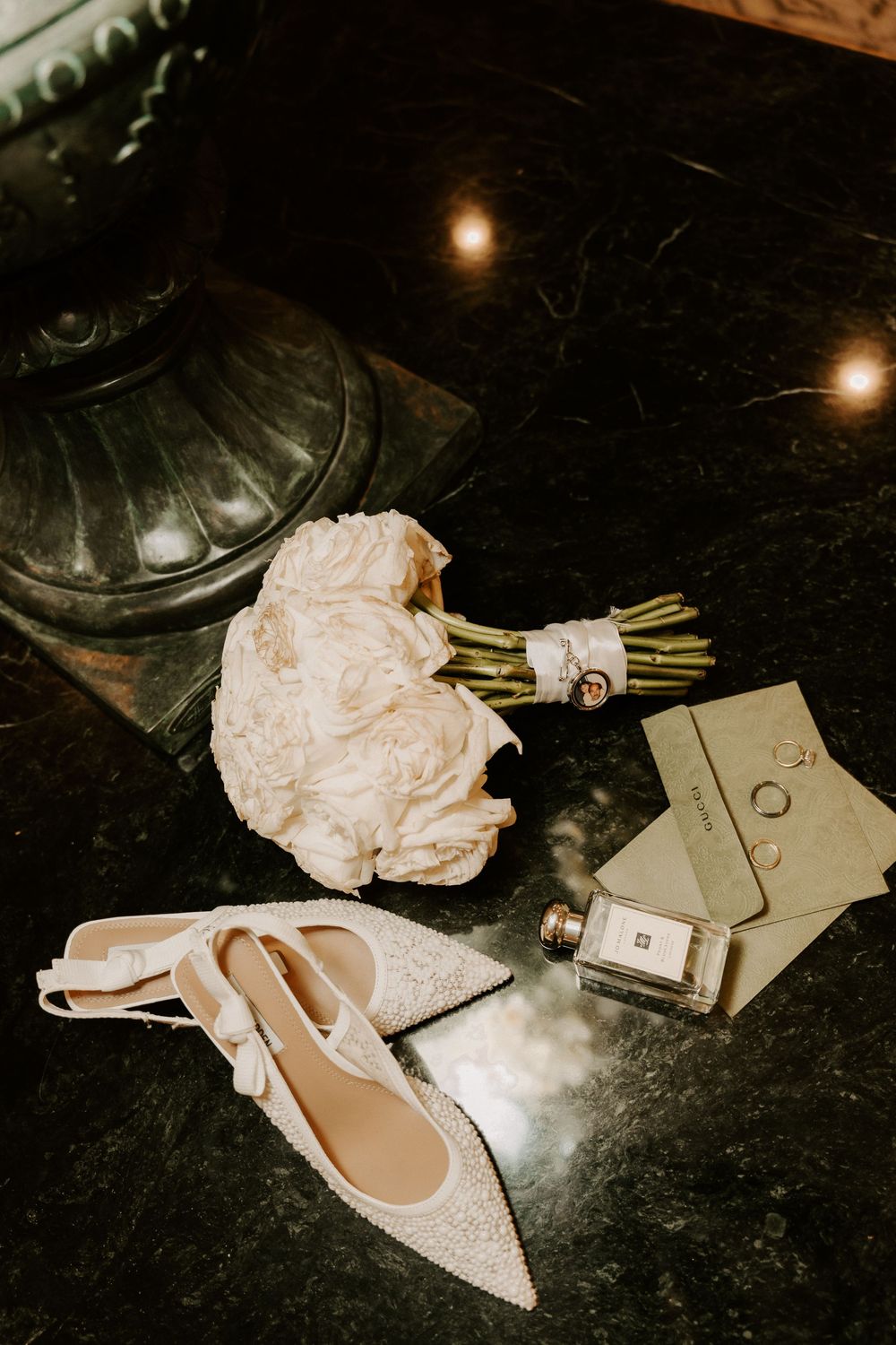 Elegant bridal flat lay featuring a white peony bouquet, pearl embellished heels, perfume bottle, and jewelry on dark marble.