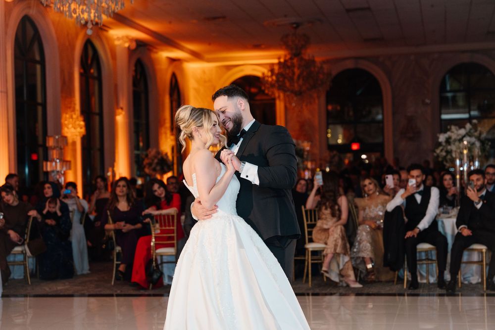Bride and groom share their first dance on a marble floor surrounded by wedding guests in an elegant ballroom.